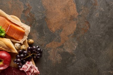 Kopya alanı ile koyu kahverengi mermer yüzeyinde çiğ balık, pastırma, üzüm, elma, baget ve bıldırcın yumurtası ile ahşap kesme tahtası üst görünümü