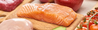 Panoramic shot of raw salmon fillet on wooden cutting board near meat and tomatoes clipart