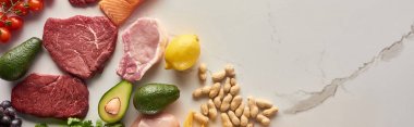 Panoramic shot of raw meat steak near avocados, lemon, peanuts on marble surface with copy space clipart