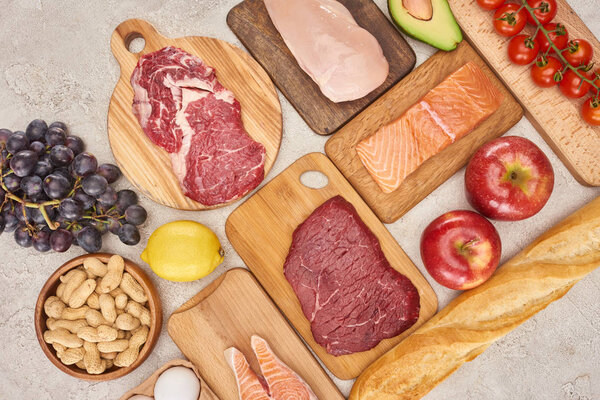 Вид сверху на сырое органическое мясо, птицу, рыбу, яблоки, виноград, лимон, авокадо, помидоры и арахис на деревянных шкафах со свежим багетом на мраморной поверхности
