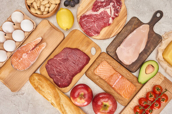 Top view af raw organic assorted meat, poultry, fish, eggs, apples, lemon, avocado, tomatoes and peanuts on wooden cupboards with fresh baguette on marble surface