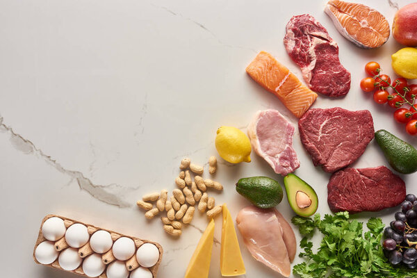 Top view of assorted meat, poultry and fish near parsley, eggs, peanuts, cheese, grapes, cherry tomatoes, avocados, apple and lemon on gray marble surface