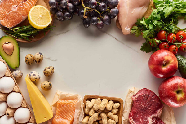 Вид сверху на разнообразное сырое мясо, птицу и рыбу с яйцами, авокадо, лимон, сыр, виноград, арахис, ветвь помидоров и зеленых веток
