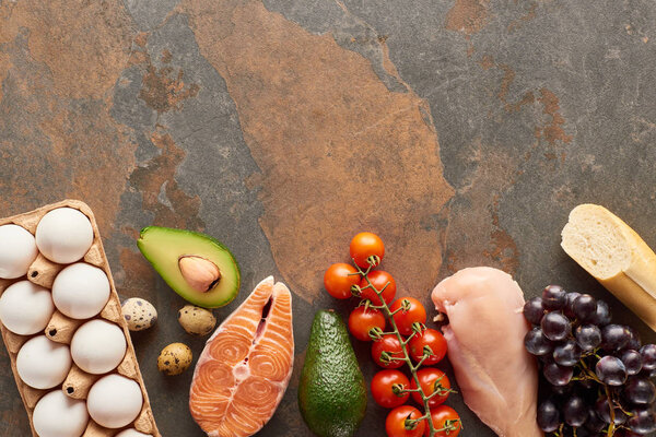 Top view of raw fish and poultry near vegetables, fruits, eggs, baguette and olive oil on marble surface with copy space