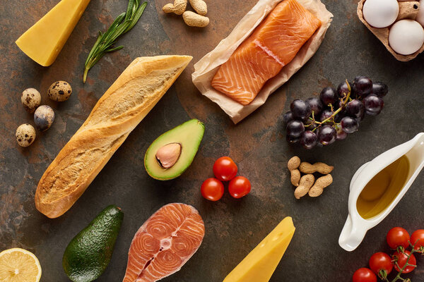 Top view of raw fish near vegetables, fruits, eggs, cheese, peanuts, baguette and olive oil on marble surface