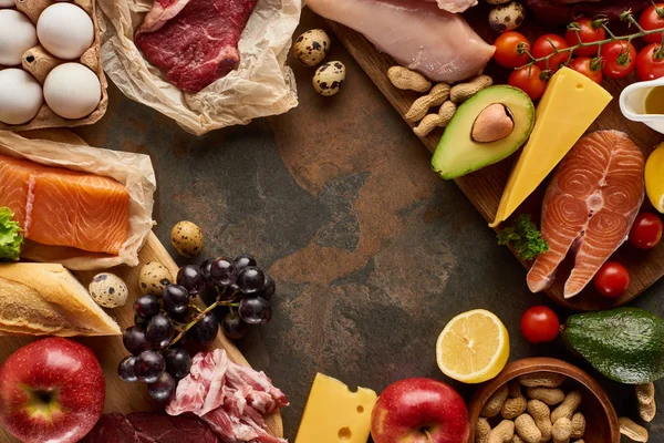 Top view of wooden cutting board with raw fish, meat, poultry, cheese, fruits, vegetables, olive oil, eggs, baguette and peanuts on dark brown marble surface with copy space