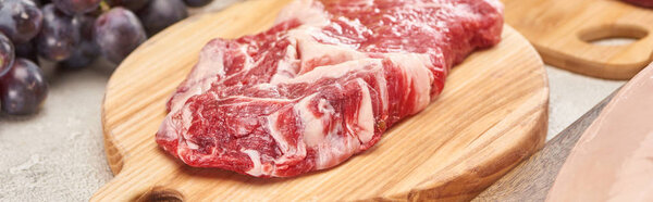 Panoramic shot of raw meat on wooden cutting board on marble surface near grape