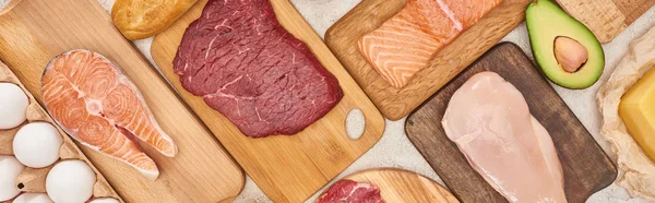 Panoramic shot raw meat, fish and poultry on wooden cutting boards near chicken eggs and half of avocado