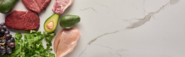 Panoramic shot of raw meat, chicken fillet, avocados, greenery and grape on marble surface with copy space