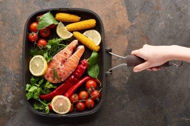 partial view of woman holding raw salmon with vegetables, lemon and herbs in grill pan clipart