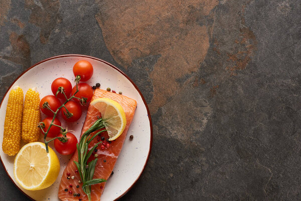 top view of uncooked salmon steak with tomatoes, corn, lemon, rosemary and pepper on plate