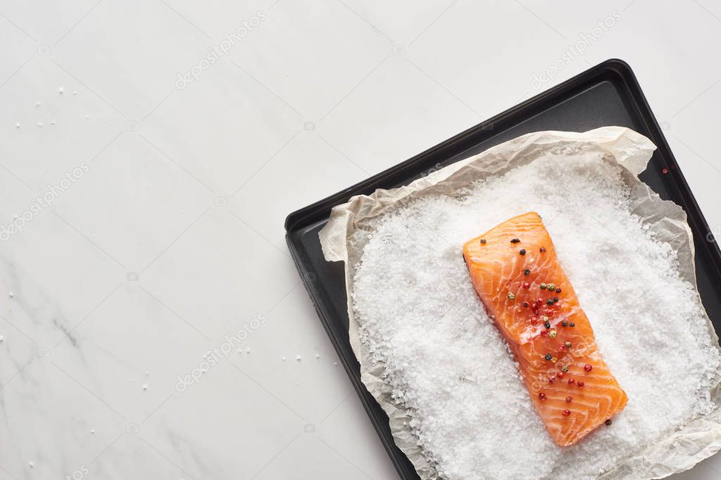 top view of raw salmon steak with pepper on oven tray with salt on marble surface