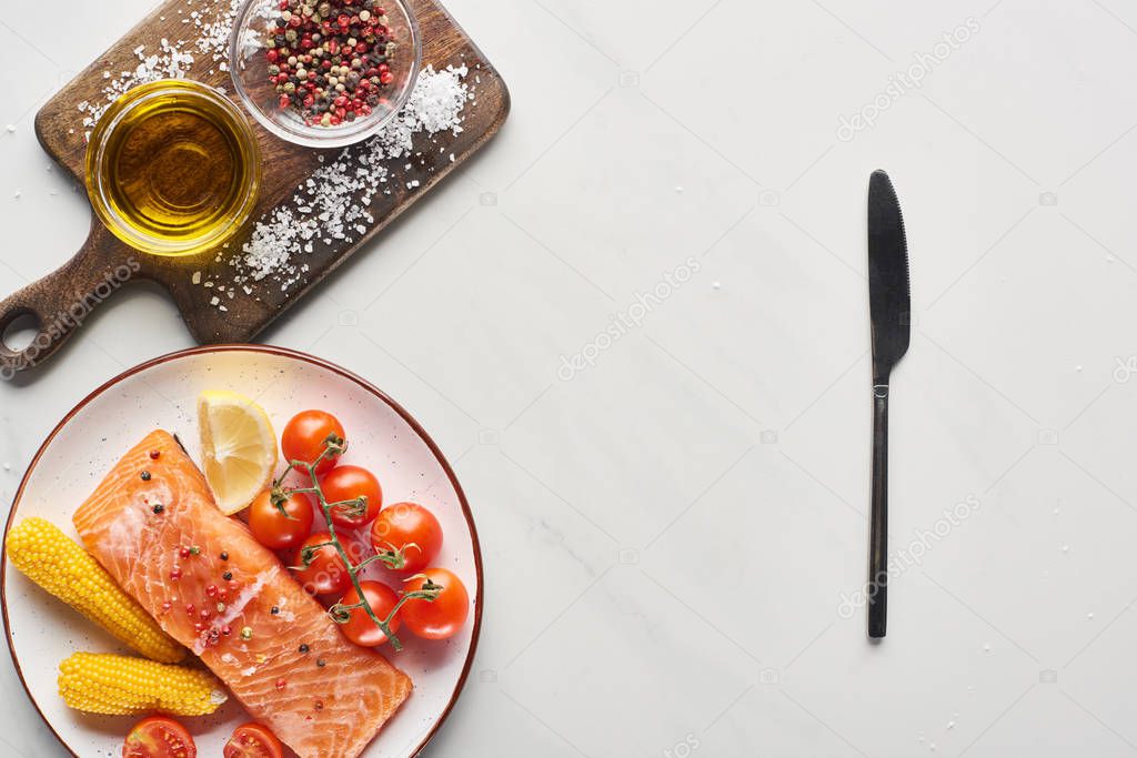 top view of raw salmon steak with corn and tomatoes on plate near knife and wooden cutting board on marble table