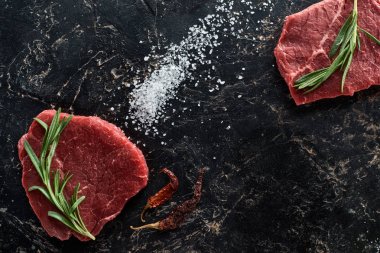 top view of raw beef steaks with rosemary twigs near dried cayenne peppers and scattered salt on black marble surface clipart