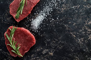 top view of raw beef steaks with rosemary twigs on black marble surface with scattered salt crystals clipart