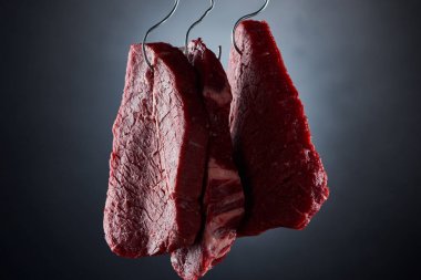 raw beef pieces on metal hooks on dark black background clipart