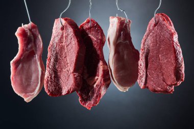 assorted raw meat steaks on metal hooks on dark black background clipart