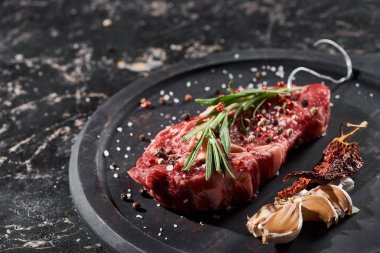 raw meat steak with rosemary twig sprinkled with salt and pepper near garlic and dried cayenne on round wooden surface clipart