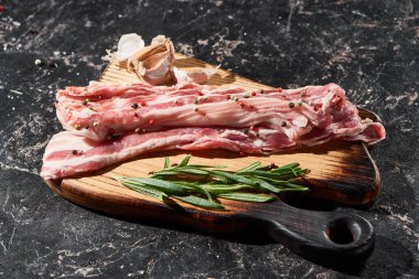 wooden cutting board with raw pork slices near rosemary and garlic on black marble surface clipart