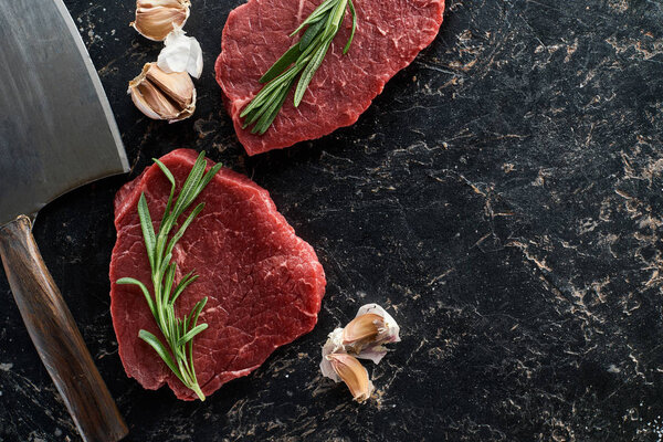 top view of raw beef steaks with rosemary twigs and garlic cloves near sharp knife on black marble surface