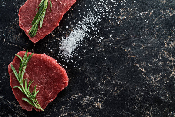 top view of raw beef steaks with rosemary twigs on black marble surface with scattered salt crystals