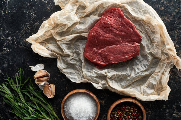 top view of raw beef steak on parchment paper near spices, garlic and rosemary on black marble surface