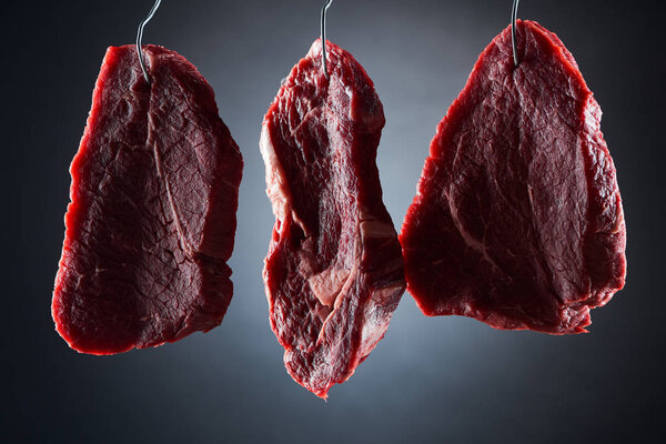 raw beef steaks on metal hooks on dark black background