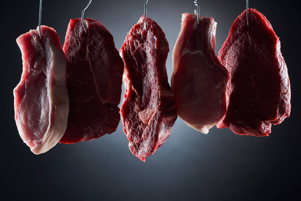 assorted raw meat parts on metal hooks on dark black background
