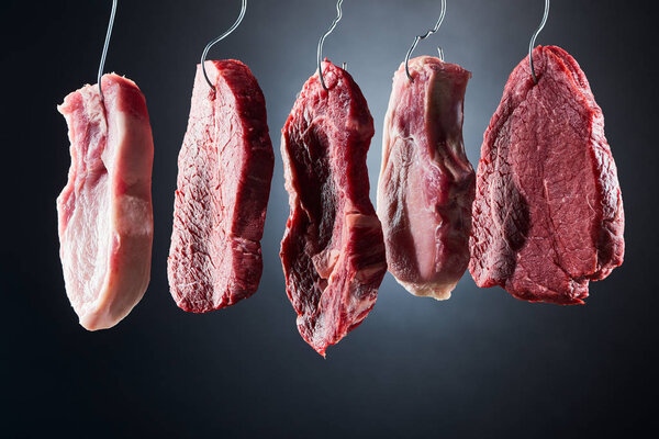 assorted raw meat on metal hooks on dark black background
