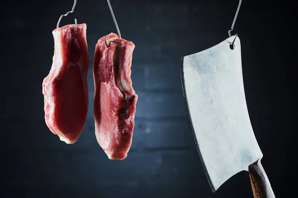 raw pork steaks and butcher knife on metal hooks on dark black background