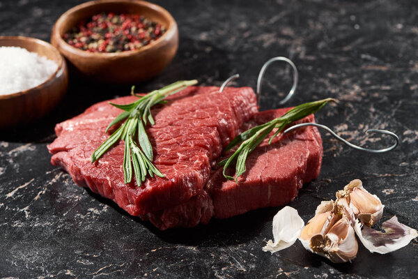 raw beef steak with rosemary near garlic cloves and small bowls with salt and peppercorns on black marble surface