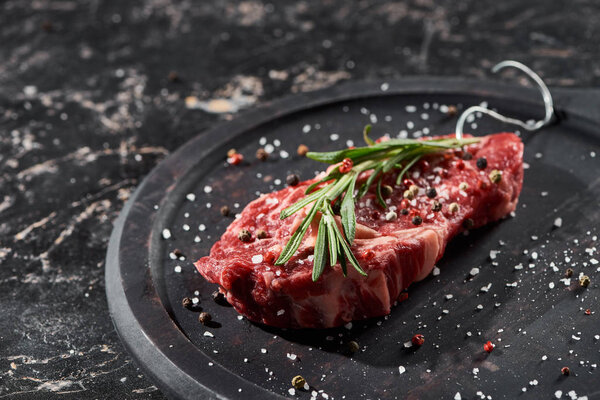 raw meat steak with rosemary twig sprinkled with salt and pepper on round wooden surface