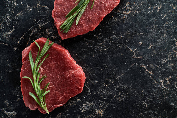 top view of uncooked beef sirloins with rosemary on black marble background