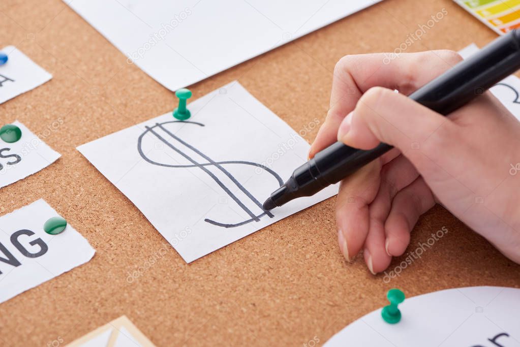 cropped view of woman writing with black highlighter dollar sign on card pinned on cork board