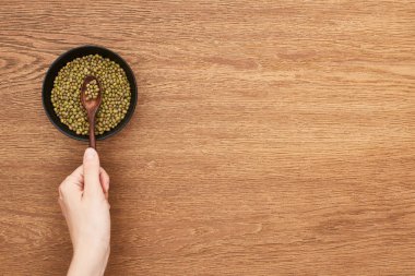 cropped view of woman holding picking with spoon raw moong beans from bowl on wooden surface clipart