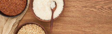 panoramic shot of bowls with white rice with spoon near roasted and raw buckwheat on wooden surface with canvas clipart