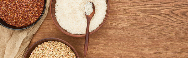 panoramic shot of bowls with white rice with spoon near roasted and raw buckwheat on wooden surface with canvas