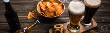 panoramic shot of glasses of light and bark beer near bottles and snacks on wooden table clipart