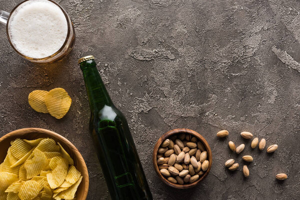 верхний вид миски с фисташками и чипсами возле бутылки и кружки пива на коричневой поверхности
