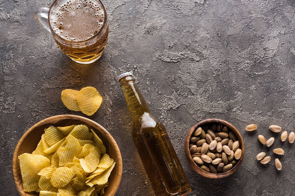 верхний вид миски с фисташками и чипсами возле бутылки и кружка светлого пива на коричневой поверхности
