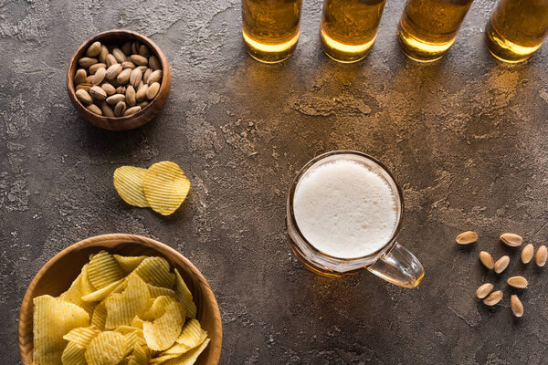 верхний вид миски с орехами и чипсами возле кружки и бутылки пива на коричневой поверхности

