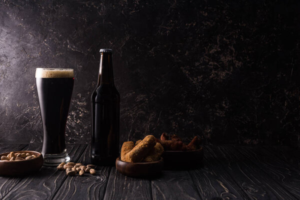 glass and bottle of beer near bowls with nuts, fried cheese and chicken wings on wooden table