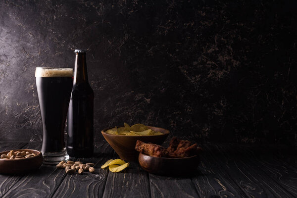 стекло и бутылка пива возле миски с орехами, чипсами и куриными крылышками на деревянном столе
