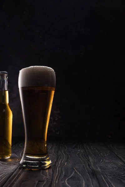glass of light beer with foam near bottle on wooden table