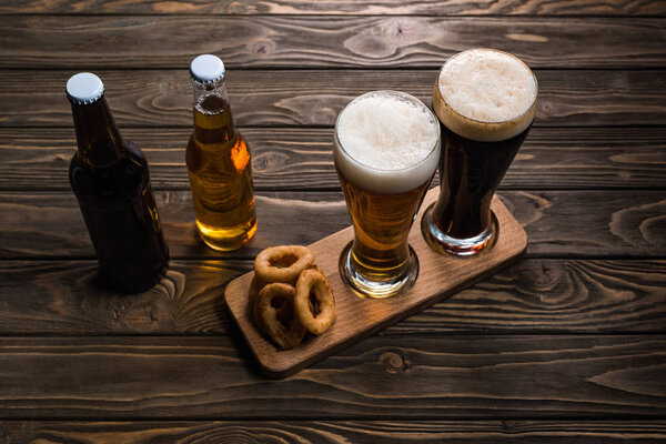 glasses and bottles of dark and light beer near fried onion rings on wooden table