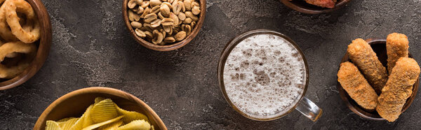 панорамный снимок кружки светлого пива возле арахиса и чипсов на коричневой текстурированной поверхности

