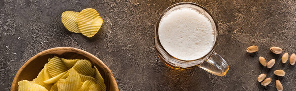 панорамный снимок кружки светлого пива возле чипсов и разбросанных фисташек на коричневой текстурированной поверхности
