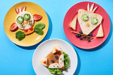 top view of plates with fancy cow, fox and bird made of food for childrens breakfast on blue background clipart