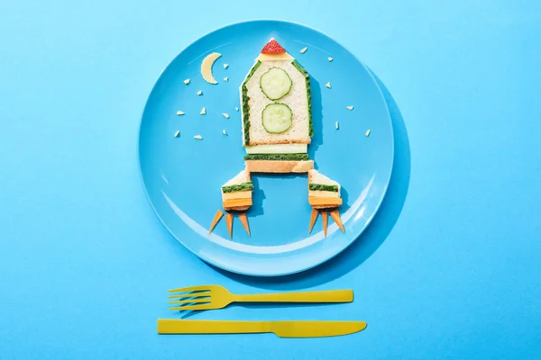 top view of plate with rocket made of food for childrens breakfast near cutlery on blue background
