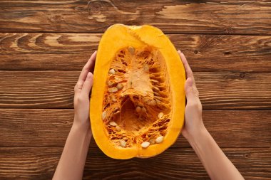 cropped view of woman holding half of pumpkin on brown wooden surface clipart